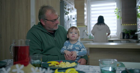 Father holding baby at dining table with colorful food and drink while mother prepares in the kitchen, warm family moment in a cozy household setting