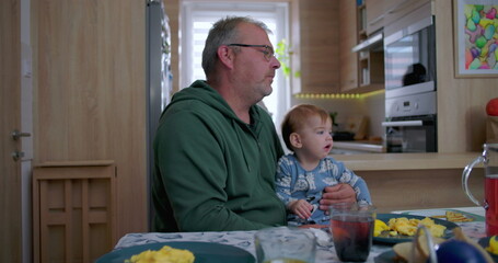 Father holding baby during family meal, baby gazing upwards with curiosity while sitting at dining...