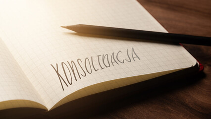 A handwritten inscription "Konsolidacja" on a grille of an open notebook on a wooden countertop, next to a black pencil, lighting of light. (selective focus), translation: Consolidation