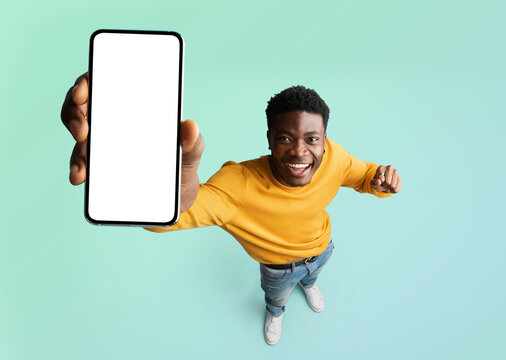 Excited black guy demonstrating smartphone with blank white screen, showing free copy space for your ad, recommending mobile app or website, standing over blue background, mockup