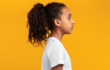 Side view profile portrait of serious black girl in white t-shirt posing isolated on yellow studio background wall and looking at empty free copy space, banner. Person with curly ponytail hair © Prostock-studio