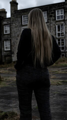 Woman facing away from old stone house, overcast day, exploring abandoned building