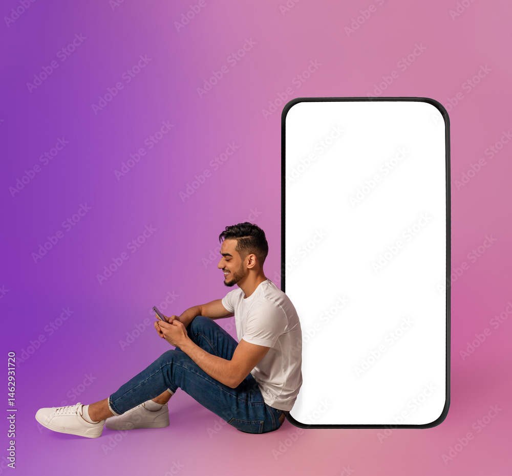 Poster cheery millennial arab guy sitting near big cellphone with blank white screen, using mobile device, 