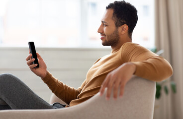 Profile side view portrait of young smiling Arab man holding mobile phone sitting on the couch in...