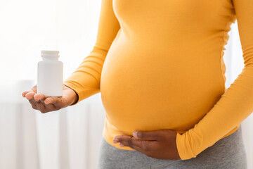 Unrecognizable black pregnant woman holding jar with pills or vitamins and touching her big tummy,...