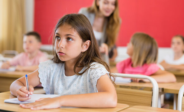 Diligent elementary school student tween girl studying with classmates, making notes of teacher lecture
