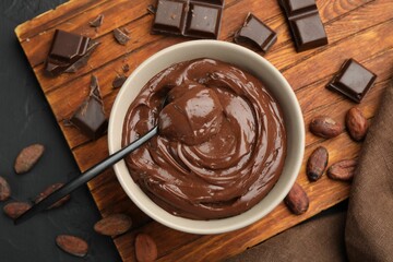 Tasty pudding in bowl, pieces of chocolate and cocoa beans on black table, flat lay