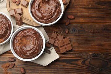Tasty pudding in bowls, pieces of chocolate and cocoa beans on wooden table, flat lay. Space for text