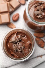 Tasty pudding in glass jars, pieces of chocolate and cocoa beans on light grey table, flat lay