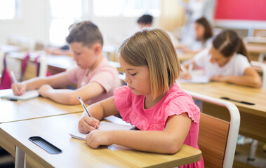 Portrait of focused cute small schoolgirl writing exercises in workbook during lesson in classroom ..