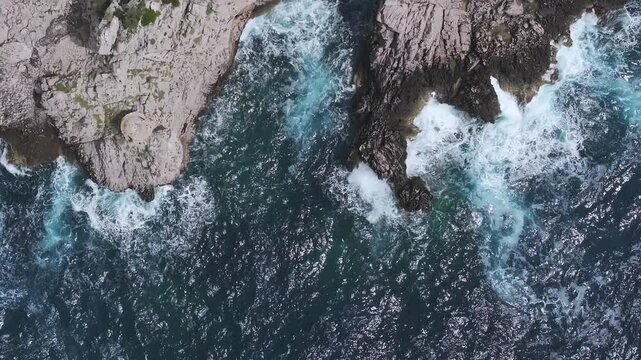 Aerial view of Capri's rugged coastline with cliffs, a natural arch, and vibrant blue waters. Waves crash dynamically near a structure with yellow rooftops.