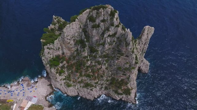 Sweeping aerial view of Capri, Italy, showcasing cliffs, rocky formations, greenery, a small beach with umbrellas, and waves crashing below.