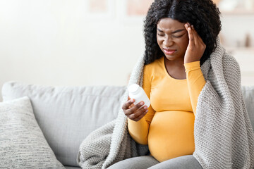 Pregnant black woman suffering from headache, taking pills to combat ache, living room interior....