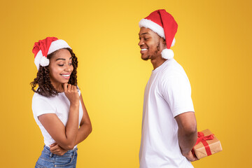Surprise For Christmas. Black Boyfriend Holding Wrapped Gift Box Behind Back Congratulating Girlfriend Standing Over Yellow Studio Background, Wearing Santa Hats. African Couple Celebrating Xmas