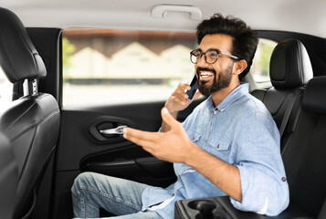 Positive young indian man talking over phone while sitting in taxi. Happy eastern guy sitting on back seat of car using mobile phone to communicate, talking over cellphone while commuting, going out