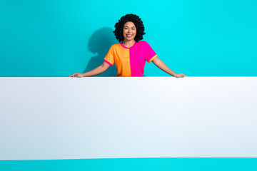 Smiling young woman in vibrant casual attire holding an empty white banner against a bright turquoise background.