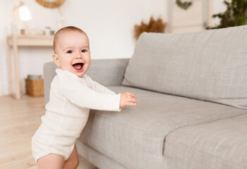 Fototapeta premium Cute Little Baby Girl Smiling To Camera Standing Near Sofa Making First Steps At Home. Happy Toddler's Portrait. Joy Of Childhood, Parenthood And Child Care Concept. Free Space For Text