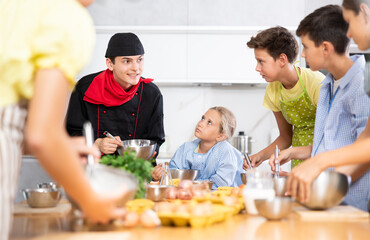 During cooking class, guy cook in black uniform stands near kitchen table with bowl in hands tells children participants culinary ticks and tricks. Man shows example of successful mobile dough
