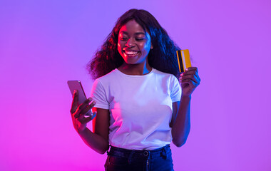 Cheerful African American young woman holding cellphone and credit card, trading or gambling...