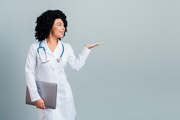Young smiling female doctor with curly hair gesturing and holding a laptop in a white coat against...