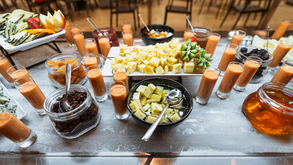 Colorful breakfast or brunch buffet table with assorted cheeses, fresh fruits, dried fruits, mini smoothies in shot glasses, jams, and honey, all beautifully arranged in a bright and elegant setting