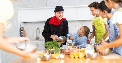 Friendly positive young guy, qualified chef, running culinary courses for group of tween children, sharing professional secrets of cooking ..