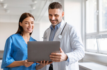 Happy male doctor and female nurse talking, working together, using laptop, discussing patient...