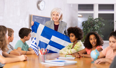 Qualified elementary school woman teacher working in a high school tells pupils the history of Greece in class and holds the .national flag of the country in her hands