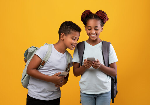 Cheerful afro-american boy and girl schoolers with backpacks using educational mobile apps on smartphones after school, black brother and sister addicted to gadgets, yellow studio background