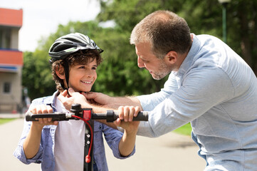 Safety Concept. Close up portrait of smiling adult father putting protection helmet on little boy at park, teaching his brave son to ride electric motorized scooter. Ourdoor Weekend Activity