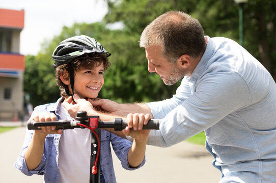 Safety Concept. Close up portrait of smiling adult father putting protection helmet on little boy at park, teaching his brave son to ride electric motorized scooter. Ourdoor Weekend Activity - Powered by Adobe