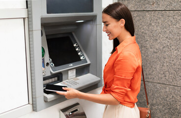 Young lady customer using smartphone to gain access to ATM, withdrawing money from account standing...