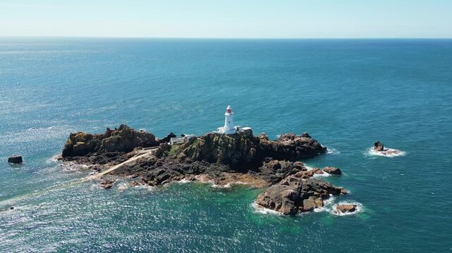 Scenic aerial drone footage of La Corbiere lighthouse. Blue sea, waves and rocky island. Jersey, Channel Islands