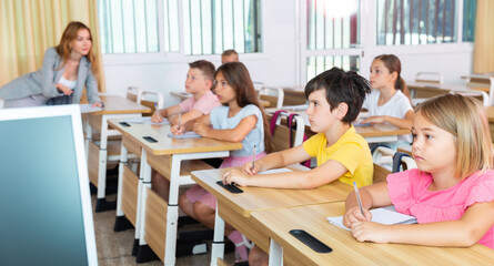 Diligent tweens studying in classroom, listening to woman teacher and writing in notebooks..