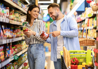 Millennial Family Couple Shopping Groceries In Supermarket Buying Food Walking With Trolley In Grocery Shop Indoor. Cheerful Customers Buy Supply Essentials Standing Near Shelves In Store Aisle