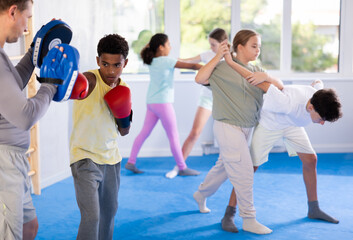 Obraz premium Motivated focused african american boy wearing boxing gloves working out with coach during group self-defense course, practicing punches on mitts..
