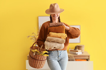Young woman in stylish hat holding warm clothes and basket with flowers near color wall at home