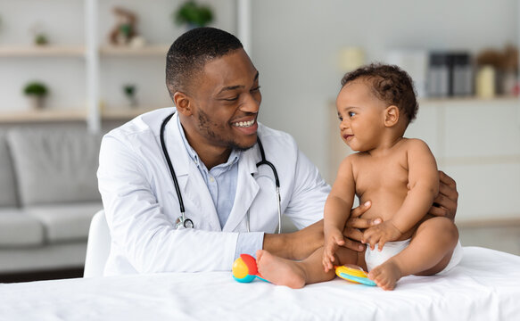 Child's Healthcare Concept. Portrait Of Smiling Black Doctor Making Check Up For Little Infant Baby Boy, Cute Toddler Child In Diaper Having Appointment At Pediatrician's Office, Free Space