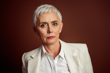 Fototapeta premium Successful mature businesswoman with short grey hair and elegant style looking poised against a minimalist brown backdrop