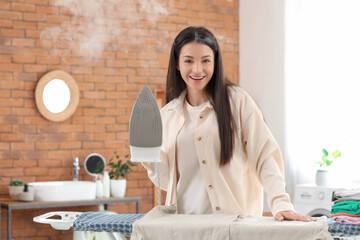 Beautiful young happy woman with iron and clothes on board in bathroom