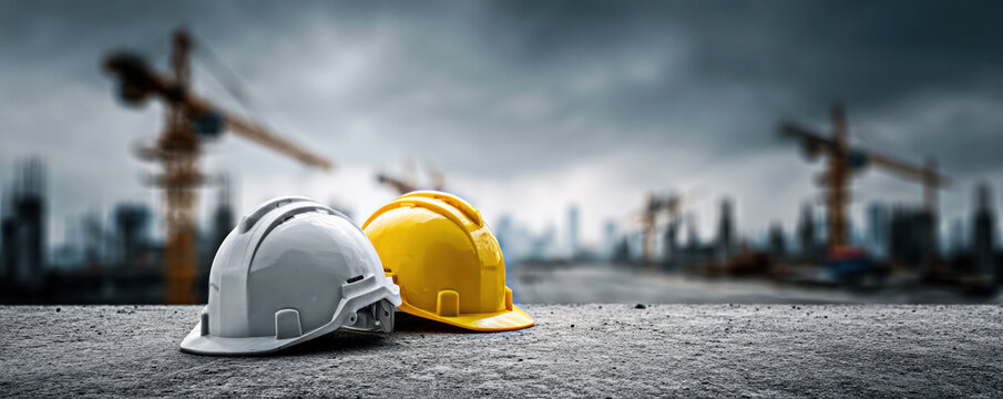Two hard hats, one yellow and one white, rest on a gritty surface in a blurred construction site background, emphasizing safety in construction and industrial environments.