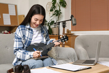 Beautiful young happy female composer with laptop writing notes while playing ukulele on sofa at...
