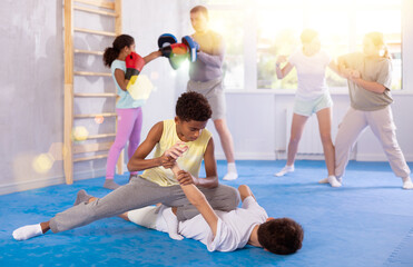 Multinational group of schoolchildren taking self-defense classes at training center. Determined african teenager applying armlock technique to face-down boy in mock fight