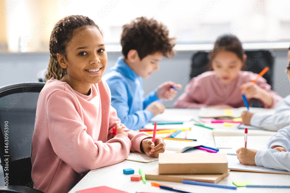 Poster academic concept. smiling junior african american school girl sitting at desk in classroom, writing 