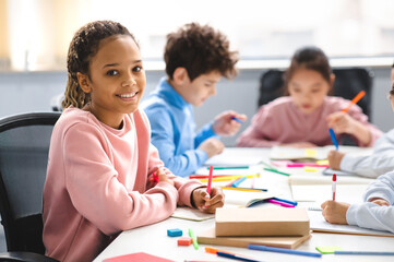 Academic Concept. Smiling junior african american school girl sitting at desk in classroom, writing...