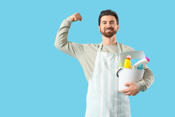 Handsome man in apron with cleaning supplies showing muscles on blue background