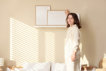 Beautiful young woman putting frame on shelf in bedroom
