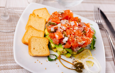 Raw salmon tartare with diced avocado sprinkled with sesame seeds served with crispy toasts, arugula, lemon and pickled capers on white plate