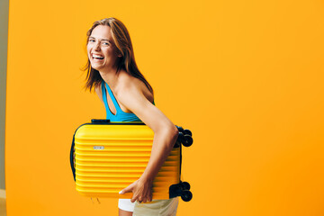 Cheerful woman holding a vibrant yellow suitcase standing against a bold orange wall with a joyful...