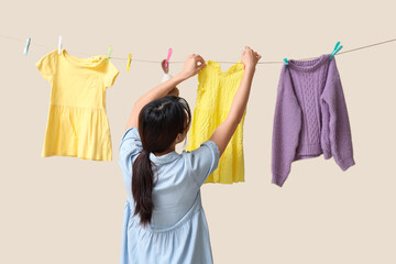 Young Asian woman hanging child clothes with clothespins on beige background, back view